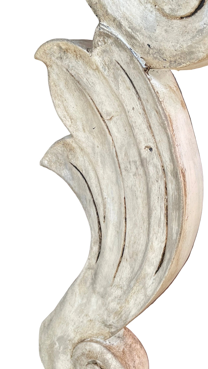 close-up of a distressed, weathered decorative wood carving with layered scrollwork and leaf-like curves in a light beige and gray tone.
