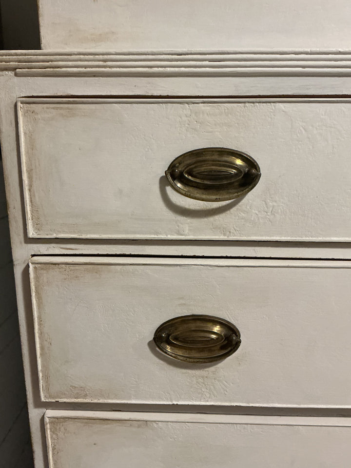 A close-up of a distressed white wooden dresser with two drawers, each featuring an oval antique brass pull handle.