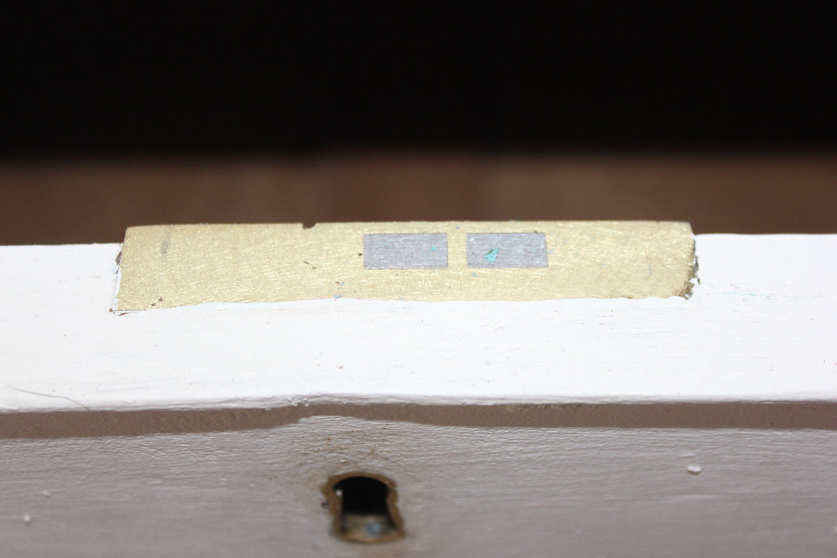 A close-up of a white painted wooden surface with a small rectangular beige strip featuring two gray squares, set against a dark background.