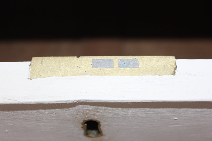A close-up of a white painted wooden surface with a small rectangular beige strip featuring two gray squares, set against a dark background.