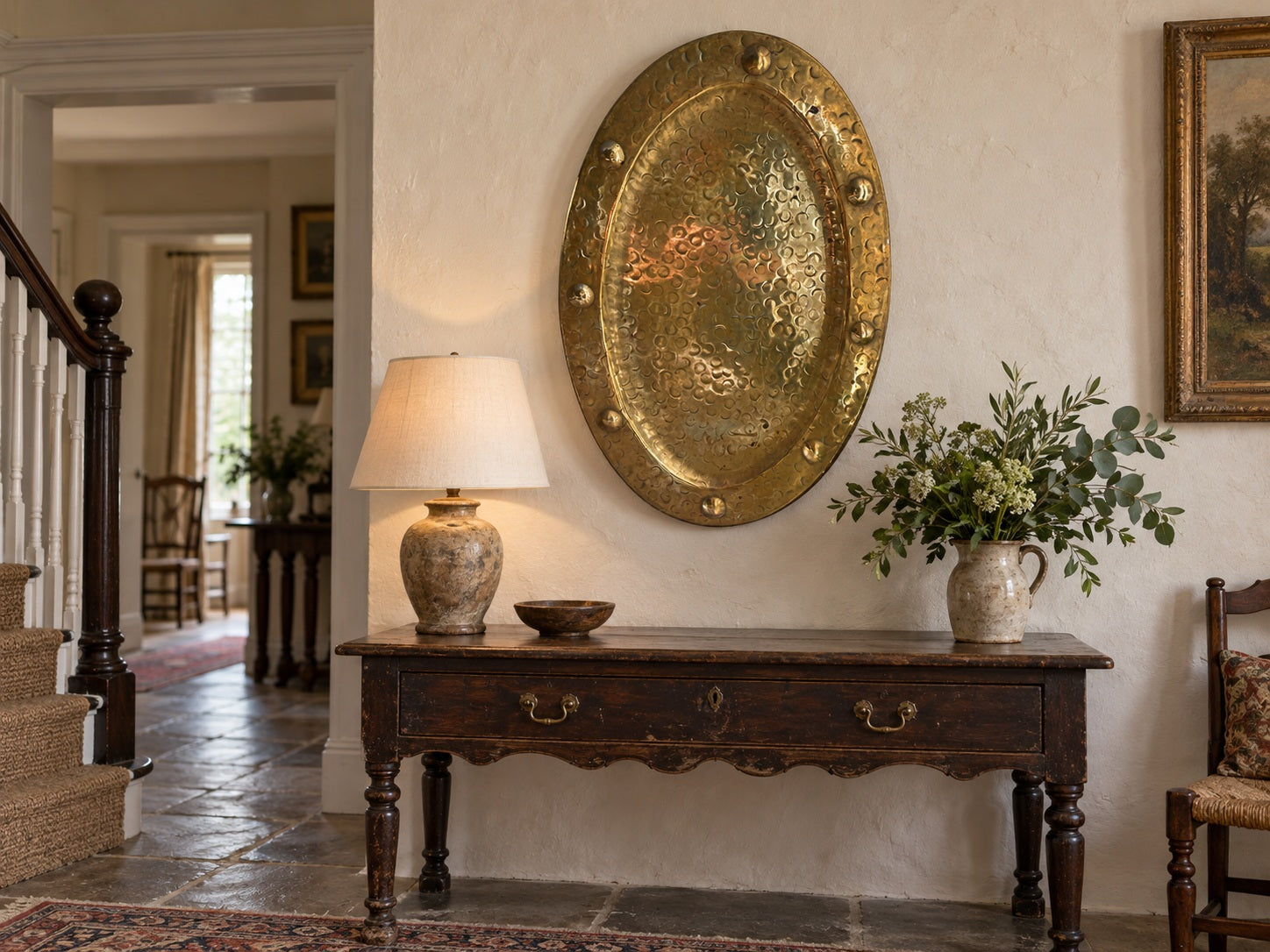 A rustic dark wood console table in a traditional home entryway, styled with a textured ceramic table lamp, a small bowl, and a ceramic vase of greenery, set against a warm cream wall with a large hammered gold oval wall hanging.

