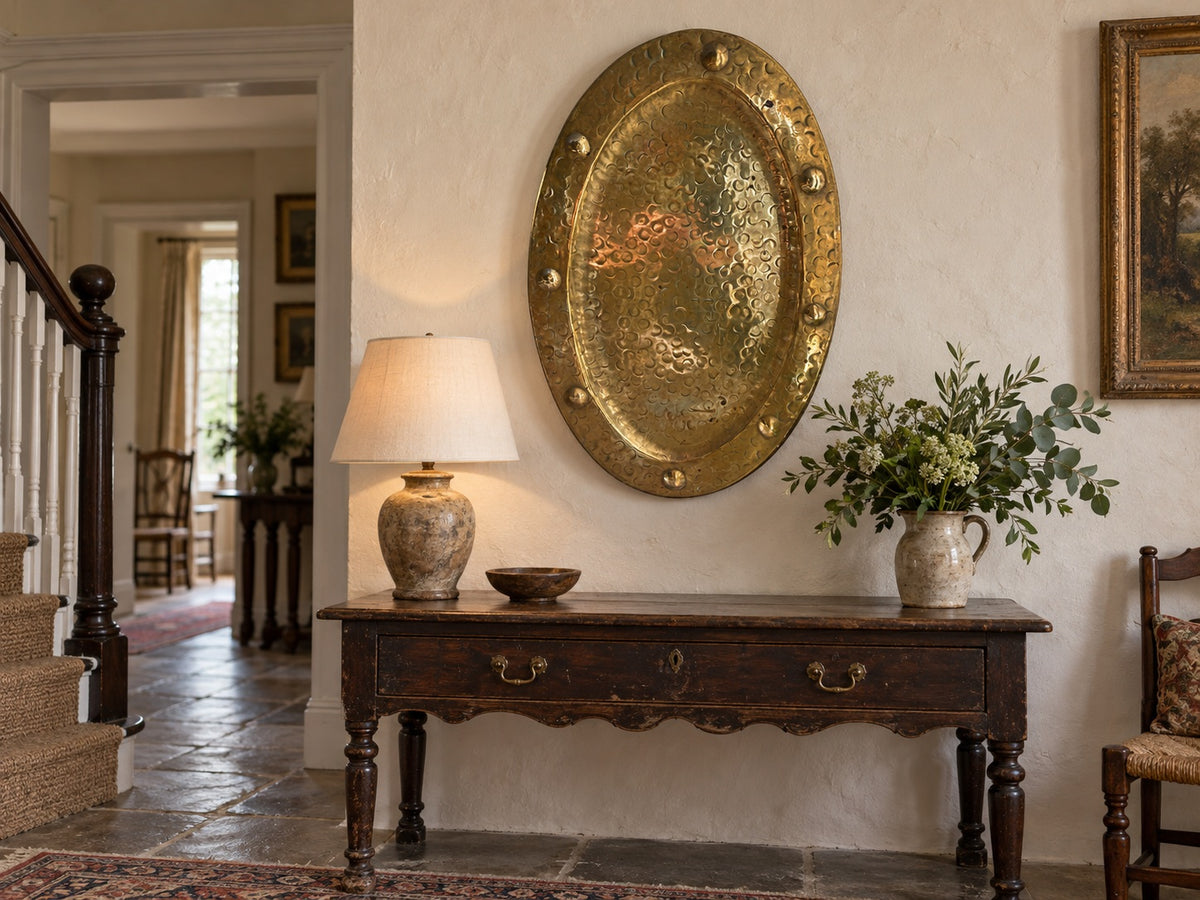 A rustic dark wood console table in a traditional home entryway, styled with a textured ceramic table lamp, a small bowl, and a ceramic vase of greenery, set against a warm cream wall with a large hammered gold oval wall hanging.

