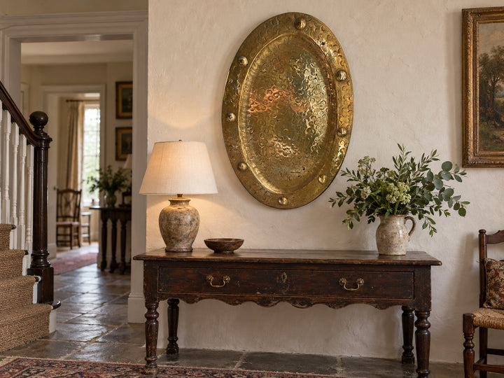 A rustic dark wood console table in a traditional home entryway, styled with a textured ceramic table lamp, a small bowl, and a ceramic vase of greenery, set against a warm cream wall with a large hammered gold oval wall hanging.

