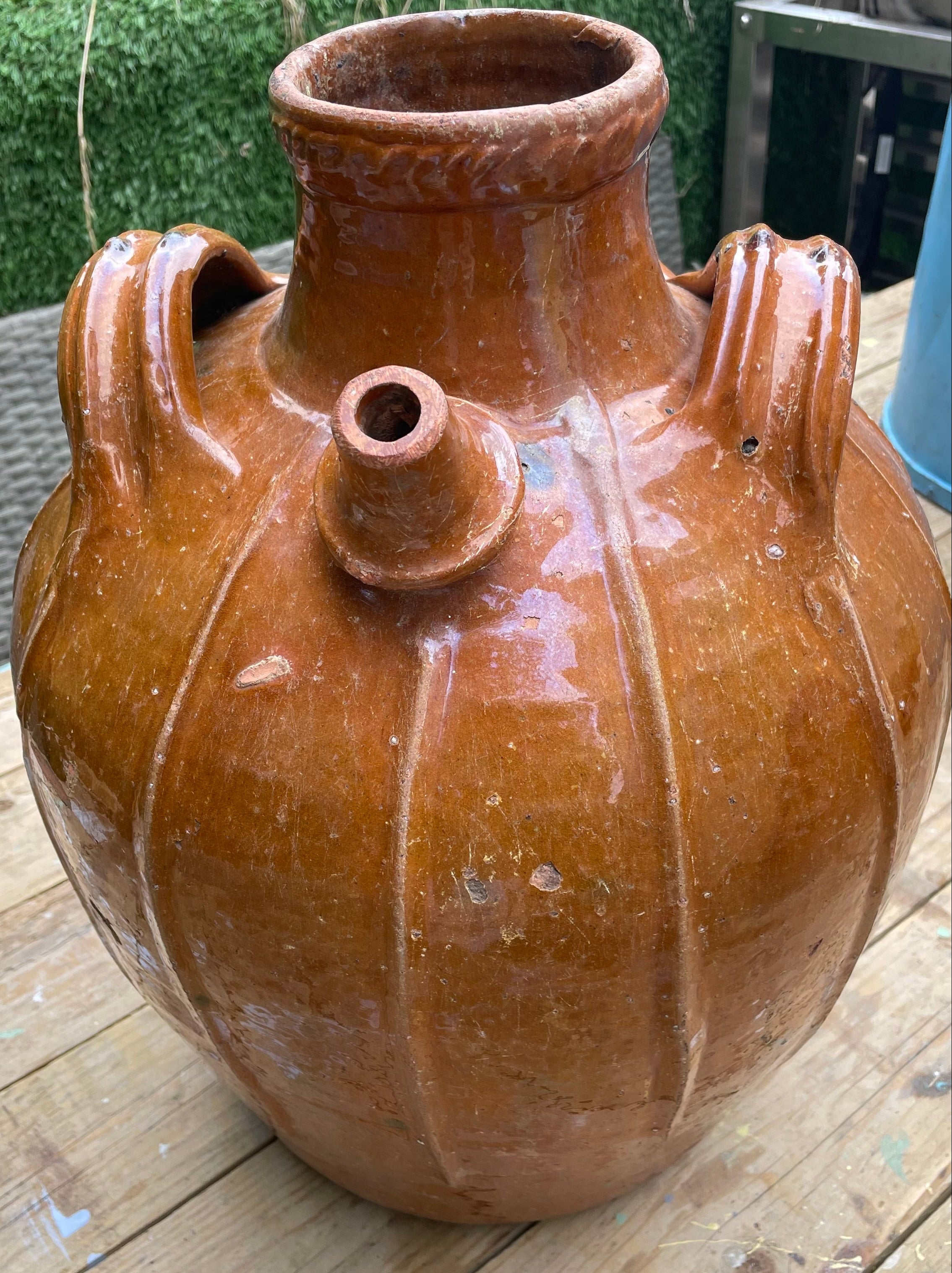 A large brown glazed ceramic vase with a ribbed pumpkin-like shape, a short neck, and two side handles, photographed on a wooden deck outdoors.