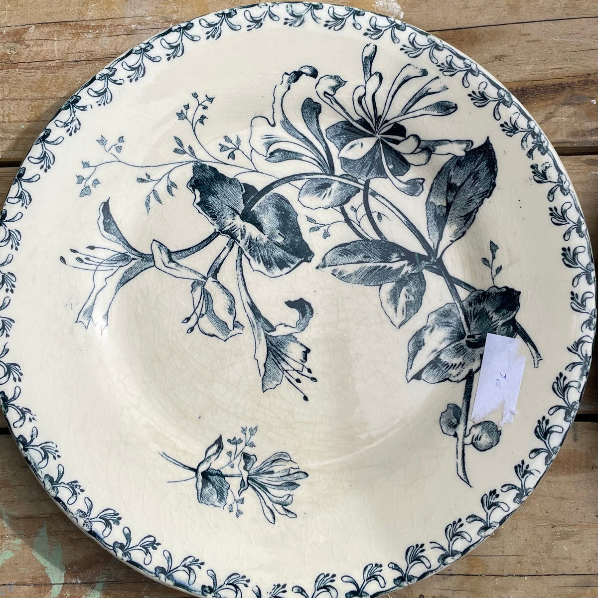 A round ceramic plate with a dark blue floral and leaf pattern on an off-white background, displayed on a rustic wooden surface.