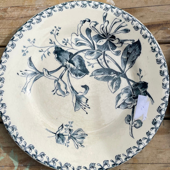 A round ceramic plate with a dark blue floral and leaf pattern on an off-white background, displayed on a rustic wooden surface.