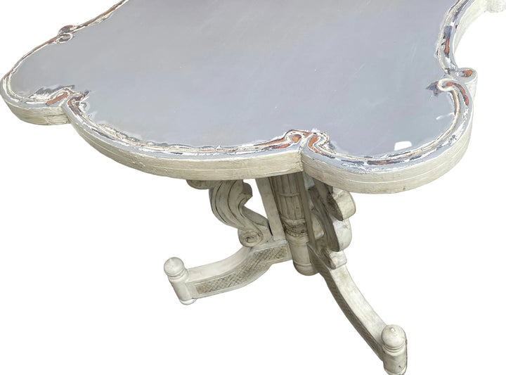 distressed white wooden pedestal table with a scalloped top, photographed outdoors on a tiled patio.
