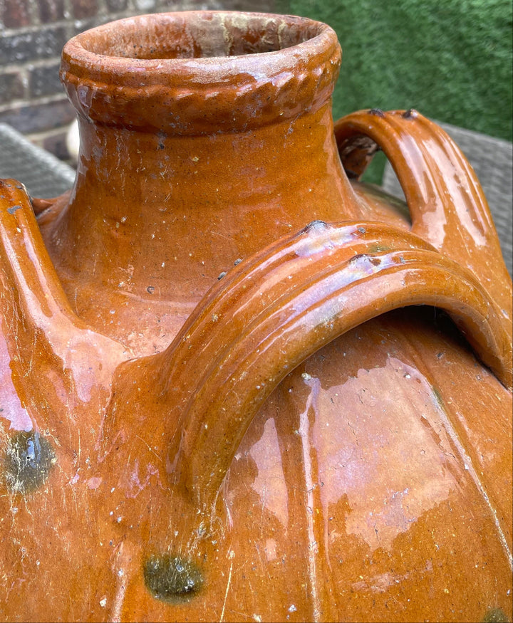 A close-up of a large glazed terracotta ceramic jug with two handles, set against a blurred outdoor garden background.