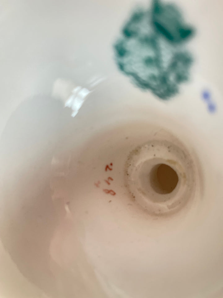 Close-up of the inside of a white ceramic bowl or cup, showing a circular rim, a small red handwritten marking, and faint green and blue painted decoration near the top.
