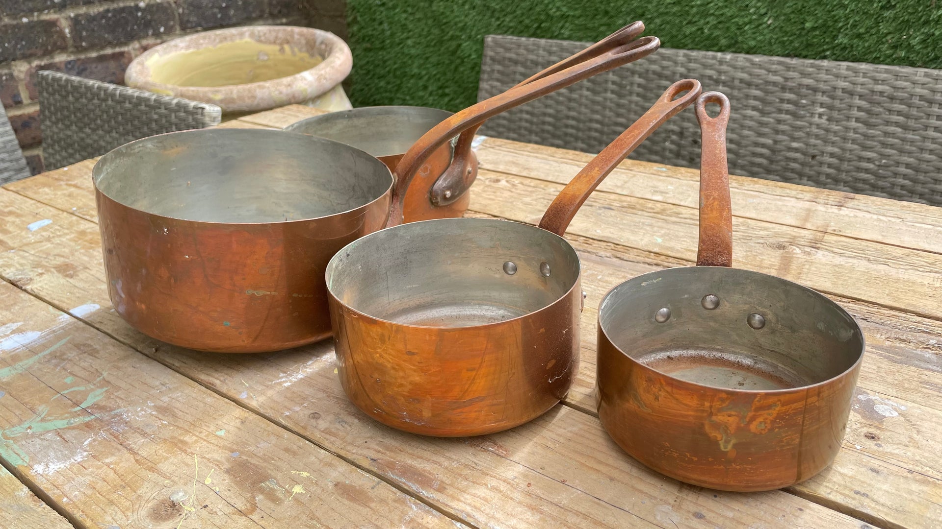 Three copper saucepans of different sizes with long handles, resting on a wooden table in an outdoor setting