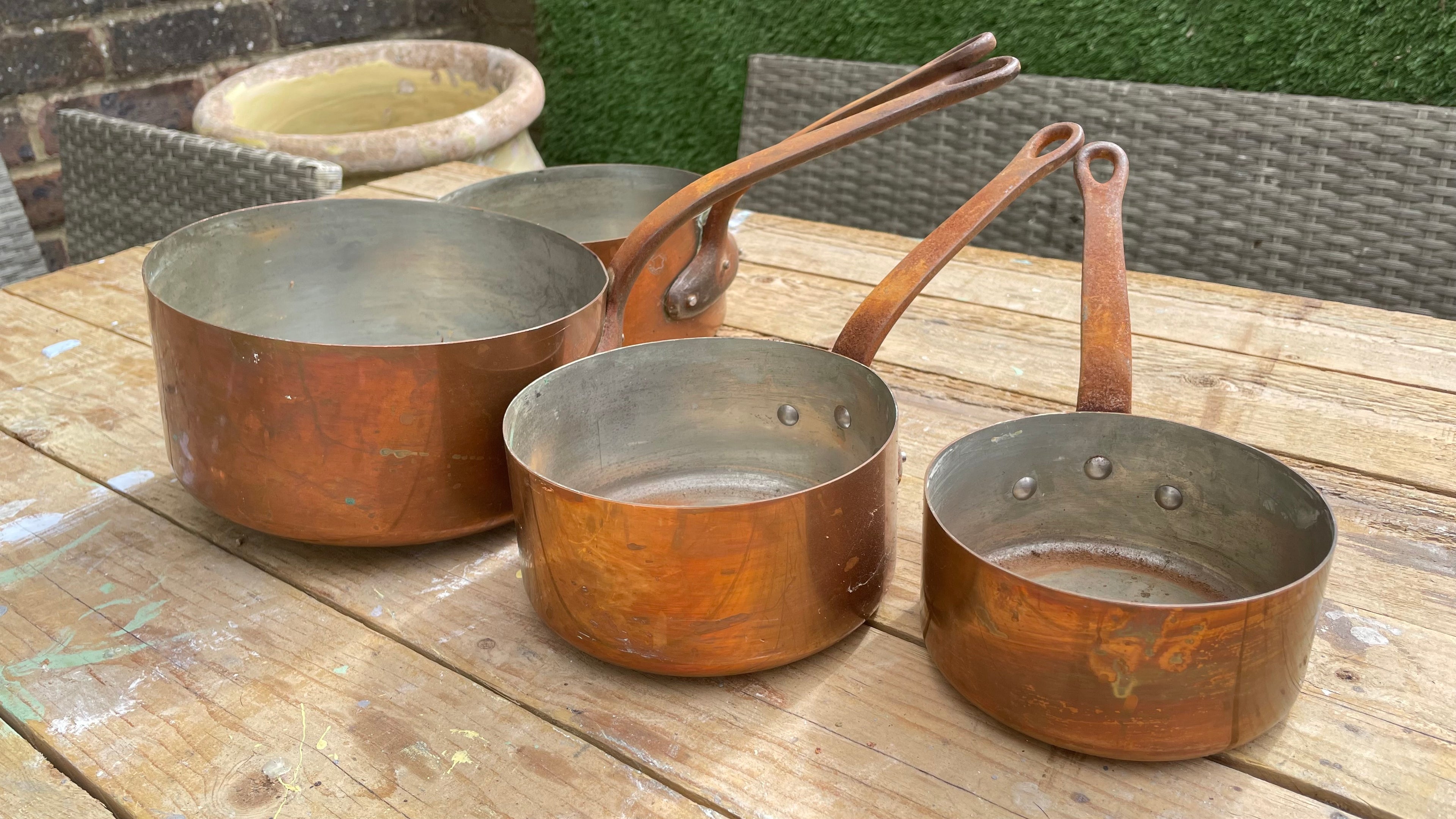 Three copper saucepans of different sizes with long handles, resting on a wooden table in an outdoor setting