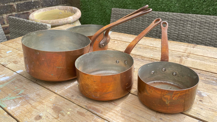 Three copper saucepans of different sizes with long handles, resting on a wooden table in an outdoor setting