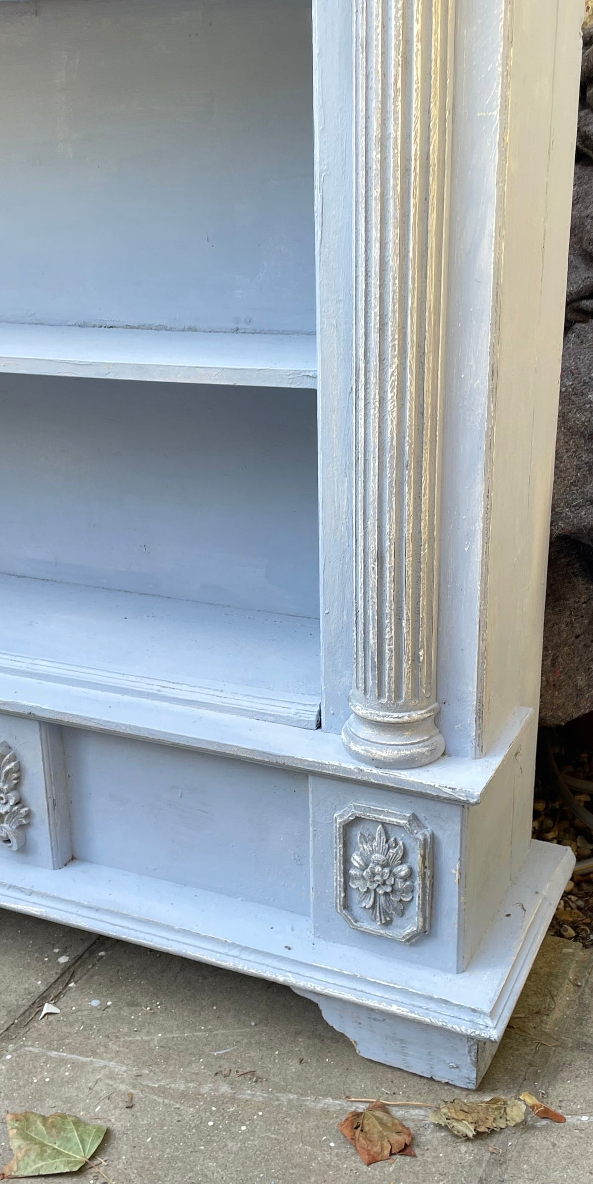 A close-up of a painted wooden cabinet or shelf unit with a light blue-gray finish and distressed silver detailing on the column and decorative panel