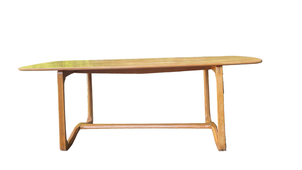 A mid-century modern wooden dining table with a warm honey finish, set outdoors on a patch of dry grass with dense green foliage in the background.