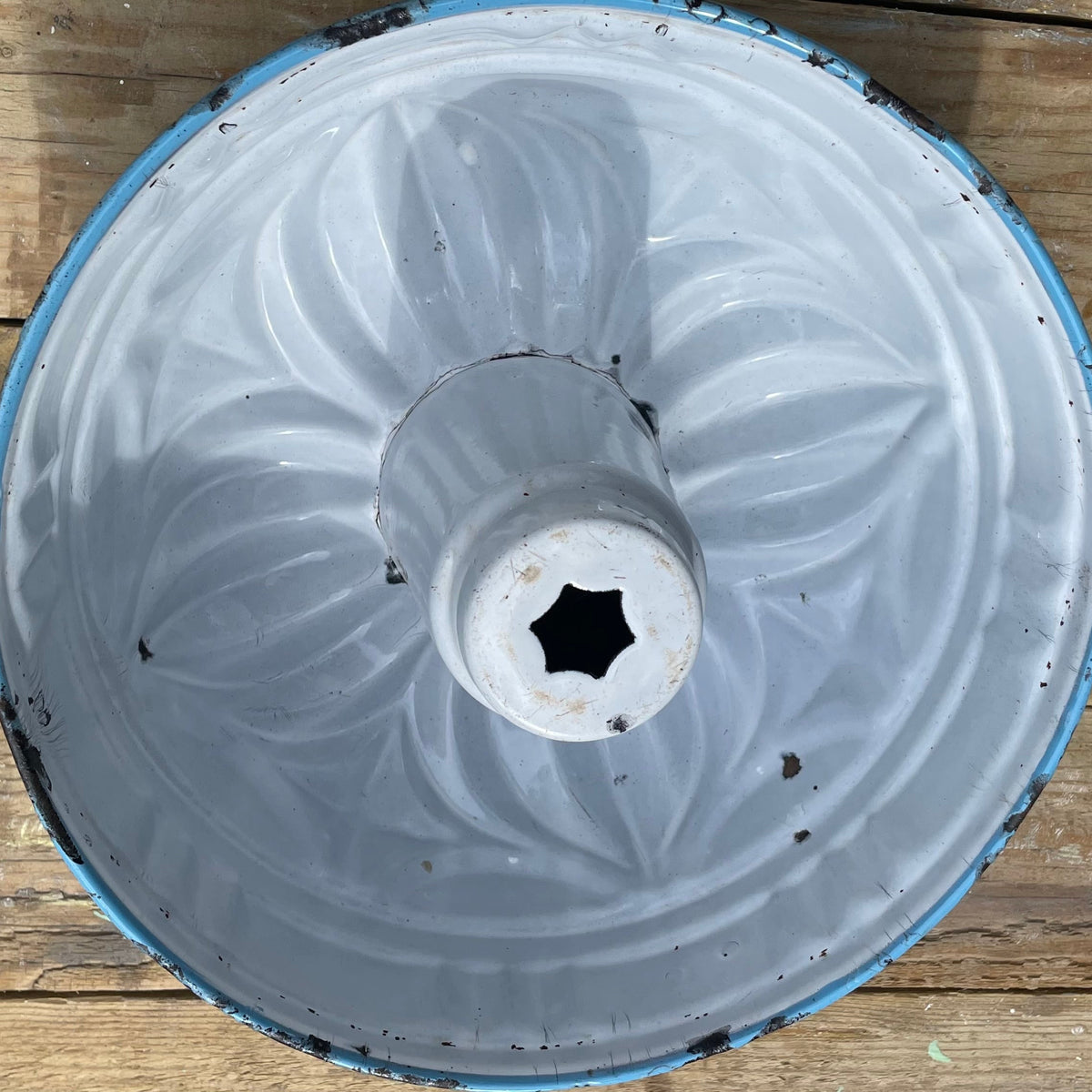A vintage round metal shade or bowl with a white enamel interior, a blue painted rim, and a star-shaped opening in the center, photographed from above on a rustic wooden plank surface.
Details