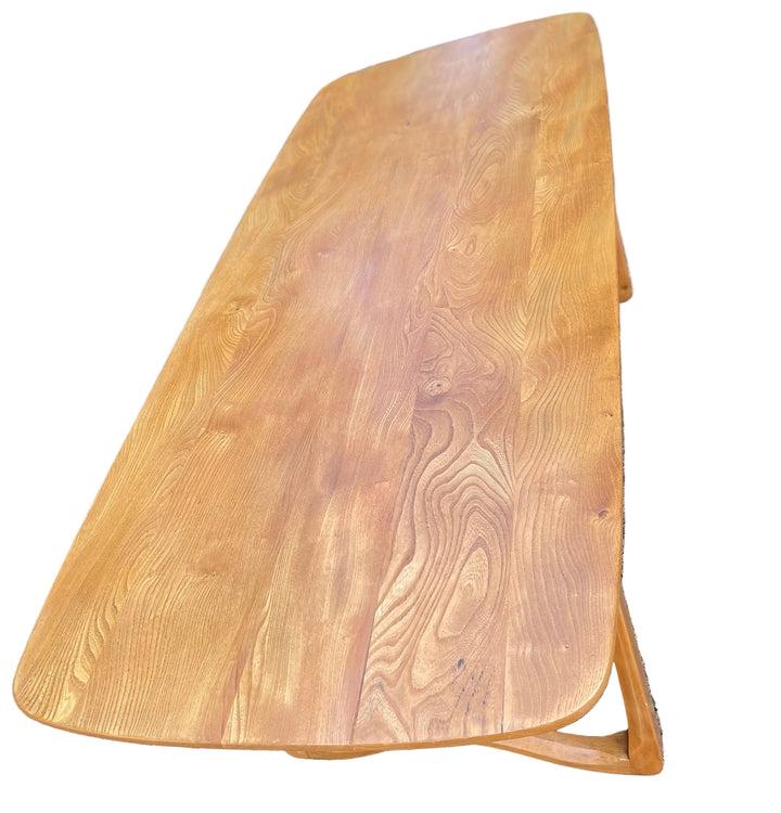 A long, rectangular wooden dining table with rounded corners and angled legs, photographed outdoors on dry grass. Sussex England.
