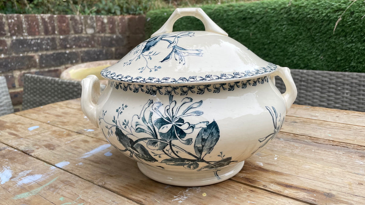 A cream-colored ceramic lidded serving tureen with blue floral illustrations, displayed on a wooden table outdoors.