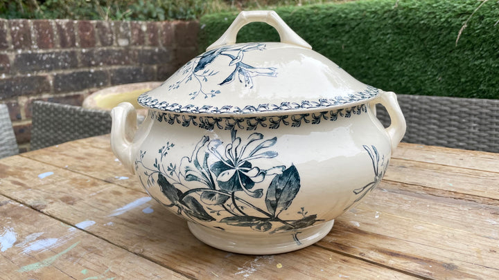 A cream-colored ceramic lidded serving tureen with blue floral illustrations, displayed on a wooden table outdoors.