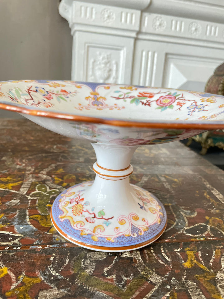 An ornate white ceramic pedestal plate with a wide rim, decorated with colorful floral and bird motifs and gold-toned detailing