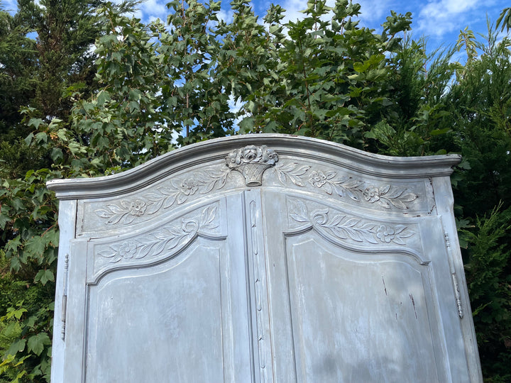 distressed light blue-gray wooden cabinet or armoire with ornate carved floral details on the top panel, photographed outdoors against a background of green trees and blue sky.