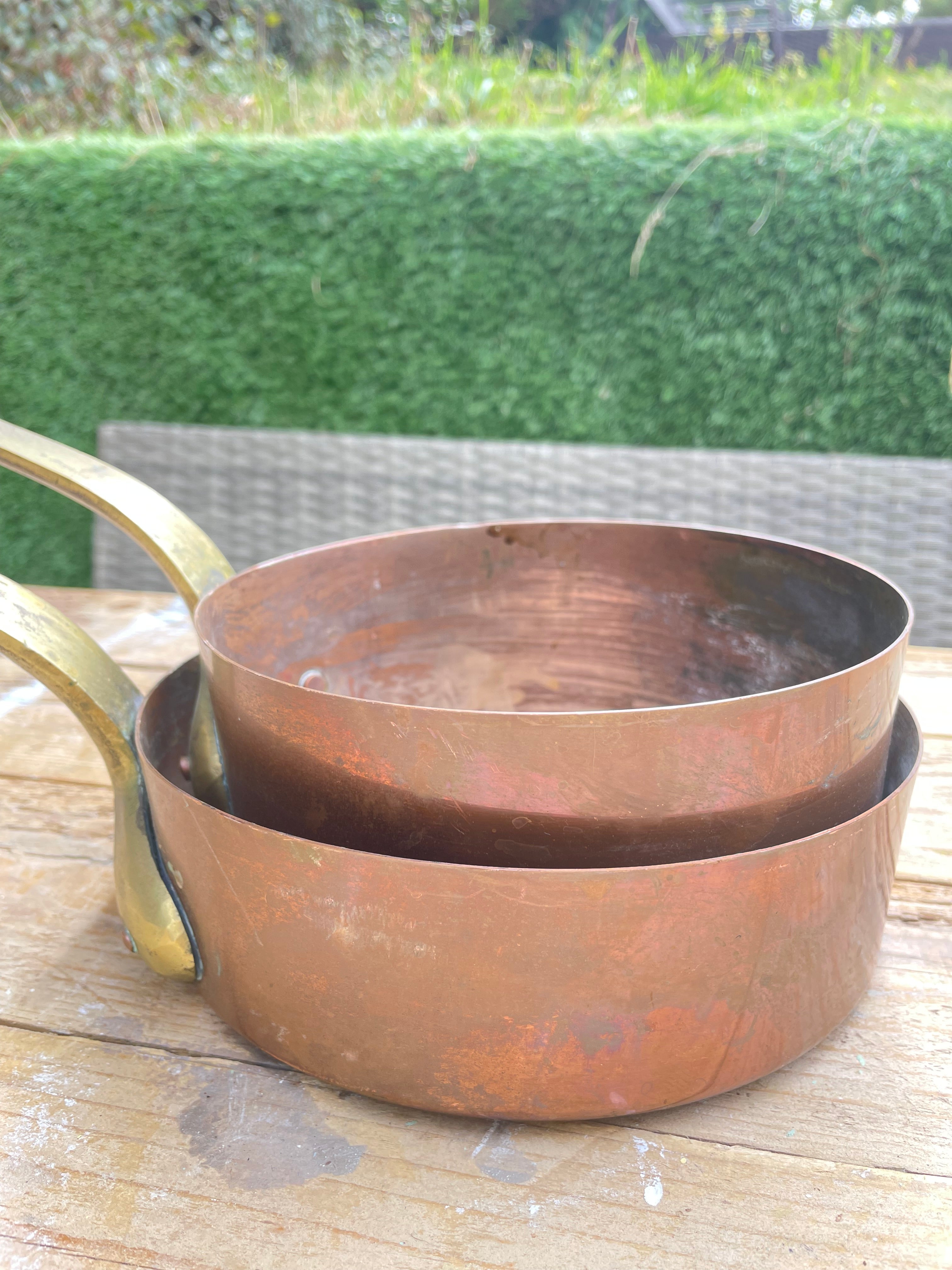 set of nested copper saucepans with brass handles resting on a wooden table outdoors, with a green hedge and patio chair blurred in the background.