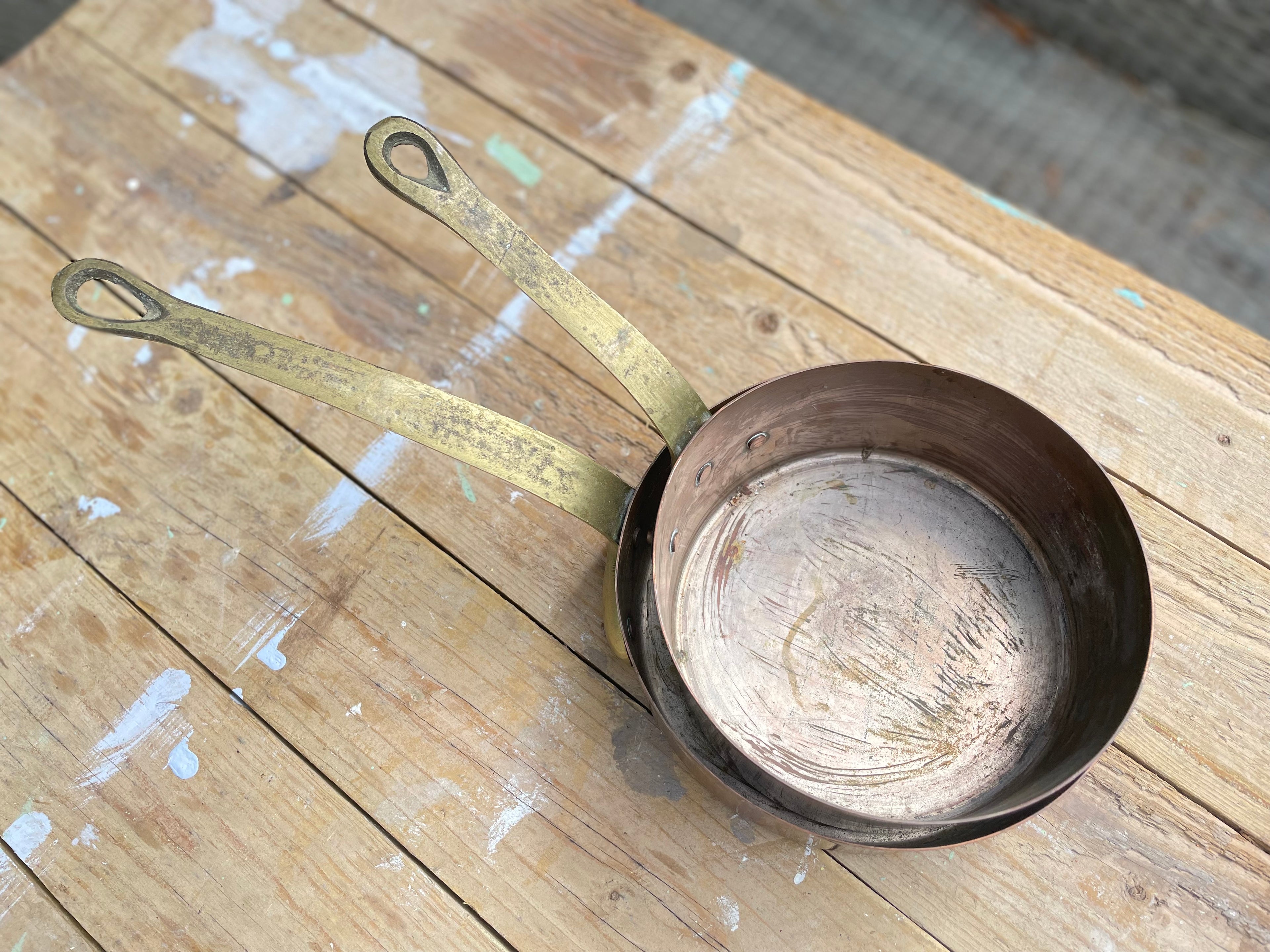 A vintage-style metal saucepan with a long, worn brass-colored handle and a dark copper-toned interior, resting on a rustic wooden plank surface.