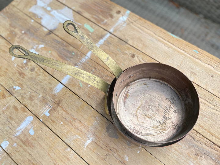 A vintage-style metal saucepan with a long, worn brass-colored handle and a dark copper-toned interior, resting on a rustic wooden plank surface.