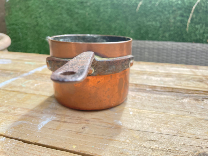 A Antique copper saucepan with a dark metal collar and a weathered wooden handle, sitting on a rustic wooden table with a green hedge in the background.
Details
