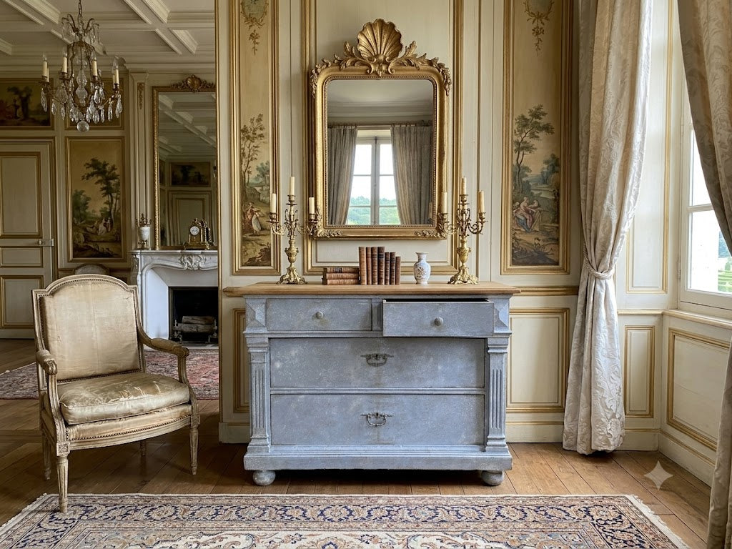 A classic French-style room featuring a distressed blue-gray wooden chest of drawers beneath an ornate gold mirror, flanked by a matching upholstered armchair and draped curtains.