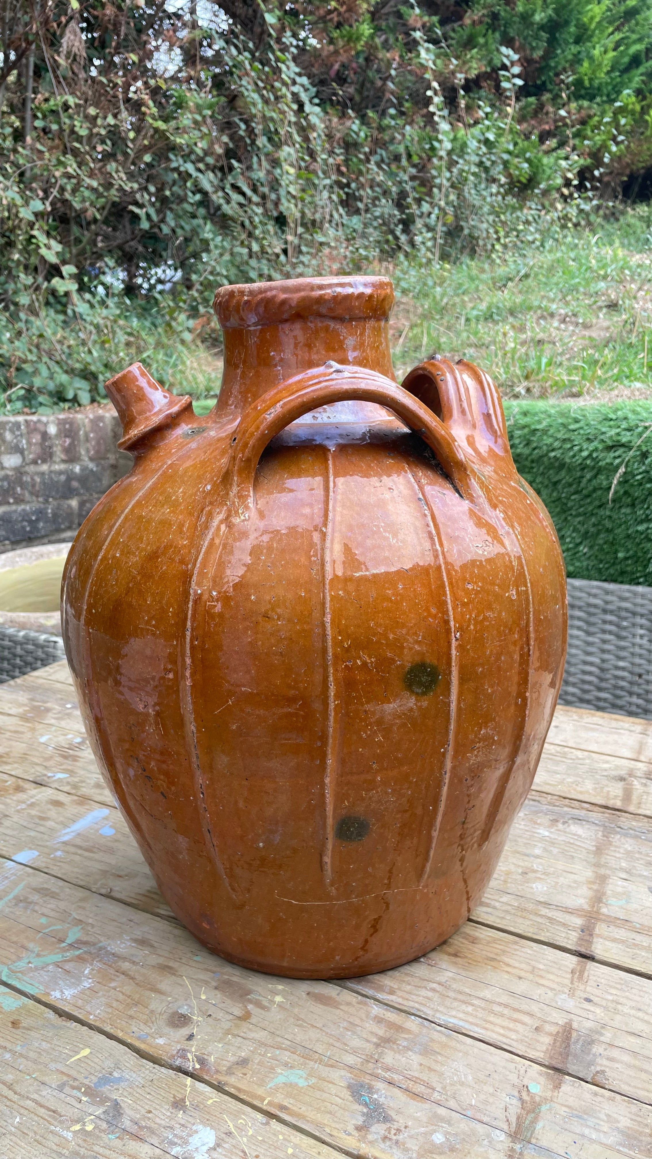 large glazed ceramic jug with a rounded body, two side handles, and a short spout, photographed outdoors on a wooden table with garden foliage in the background.
Details