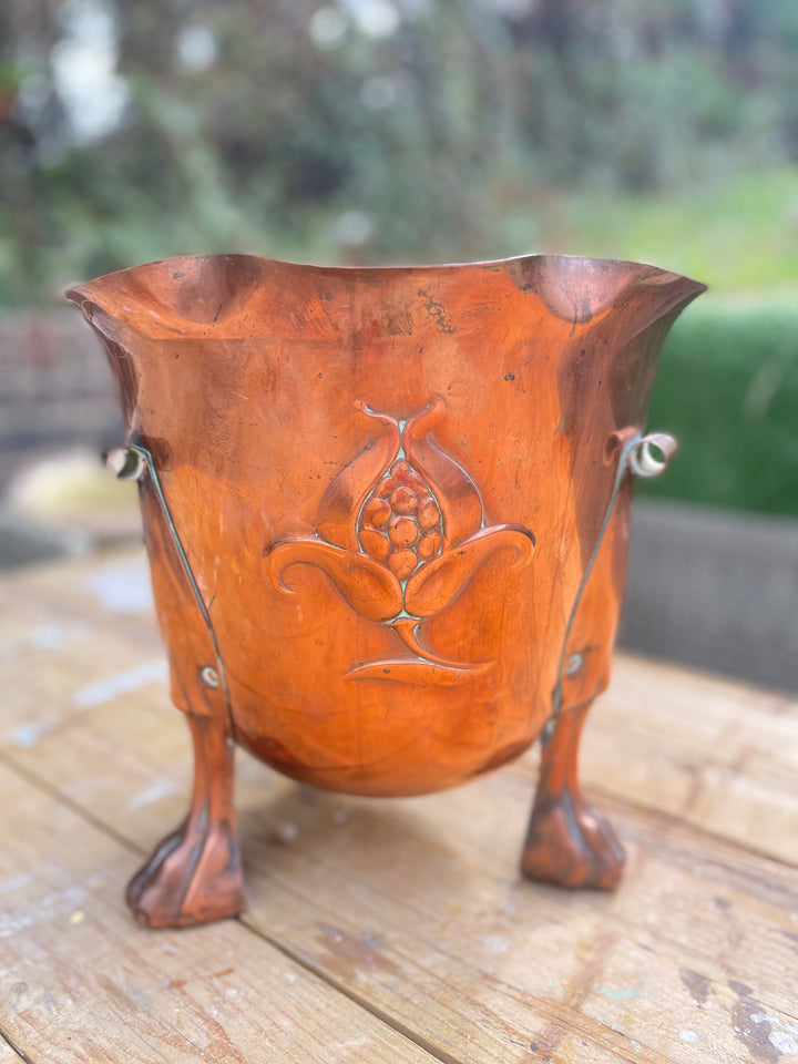A copper planter with a decorative embossed floral design and three small side handles, shown on a wooden surface with a blurred outdoor garden background.
