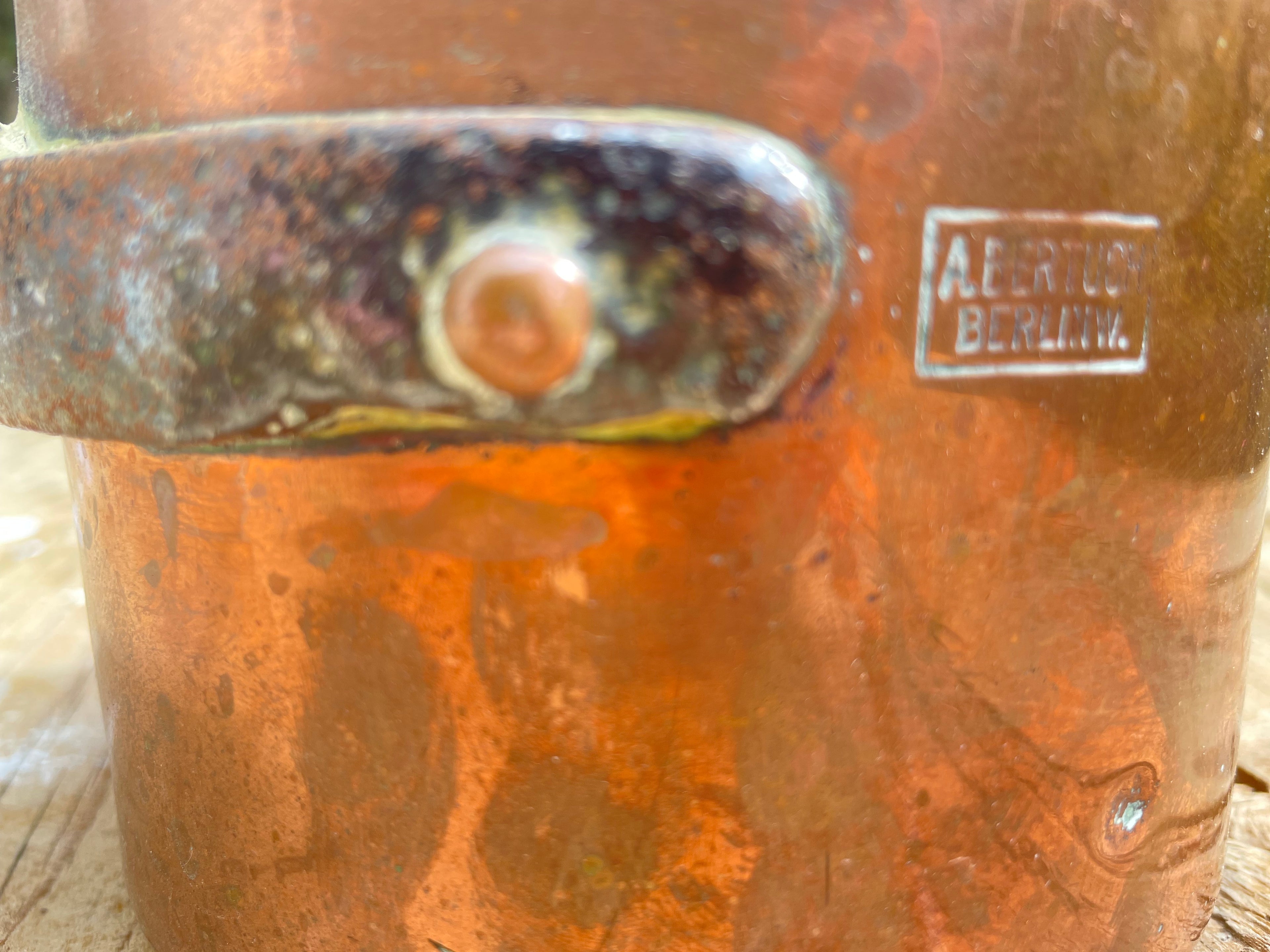 Close-up of a distressed copper-colored metal surface with a rusted handle or bracket, a round central knob, and a small rectangular metal plaque reading 'A. BERTUCK BERLIN'.