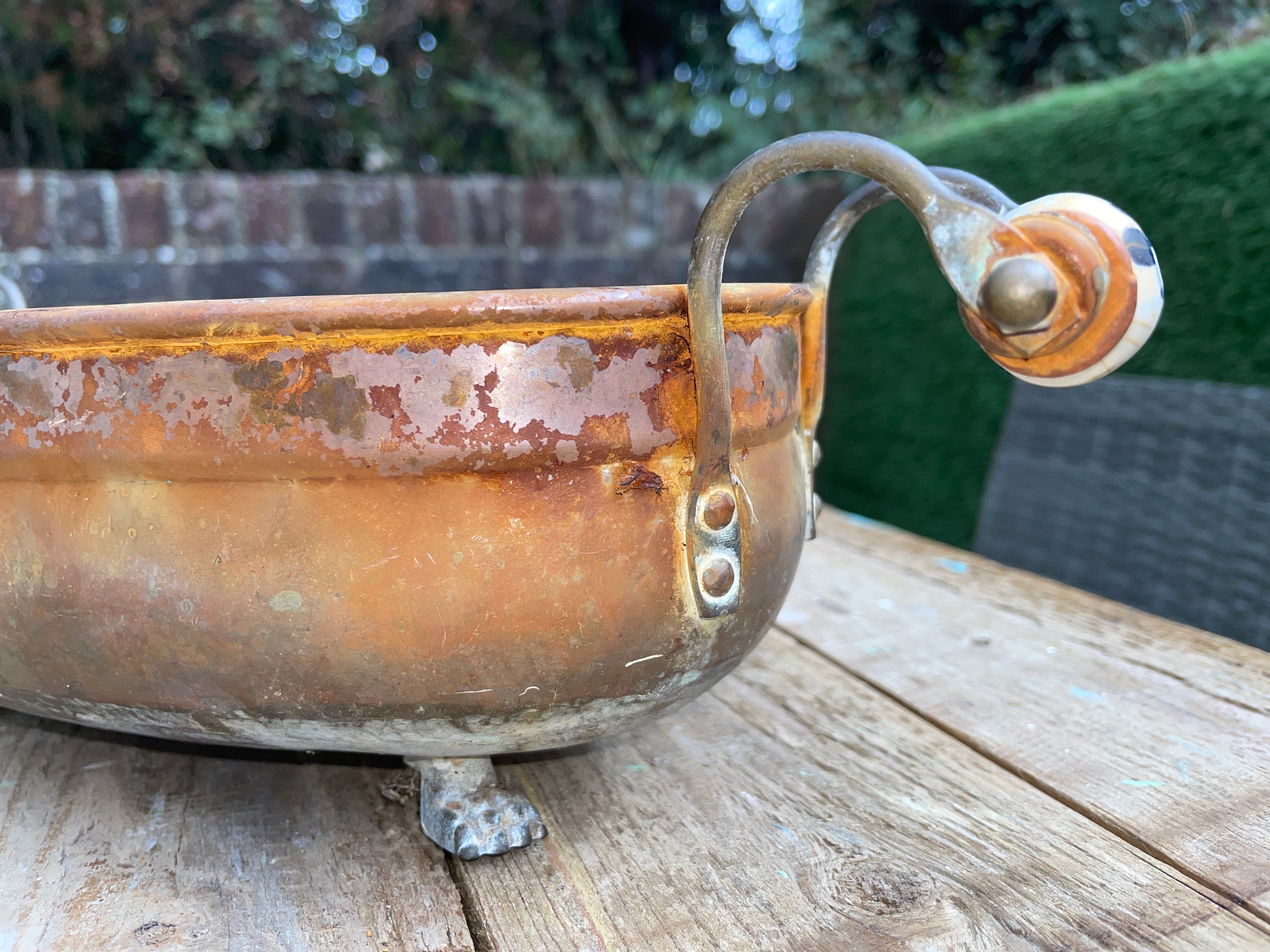 A rustic copper-colored metal bowl with a curved handle and small feet, photographed outdoors on a wooden table with a blurred garden background.
Details