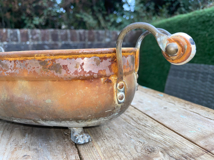 A rustic copper-colored metal bowl with a curved handle and small feet, photographed outdoors on a wooden table with a blurred garden background.
Details
