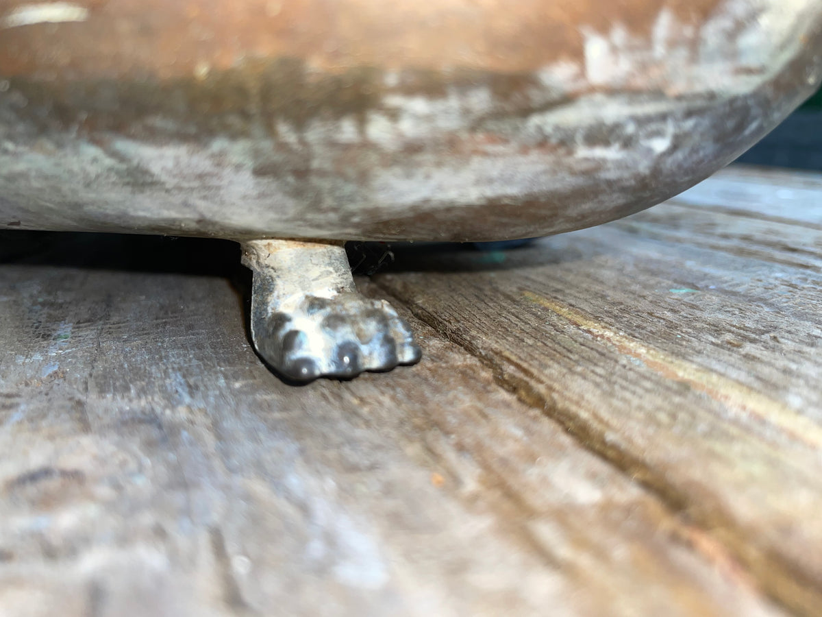 Close-up of a rustic metal bowl or planter with a small sculpted animal paw foot supporting its edge, resting on a weathered wooden surface.