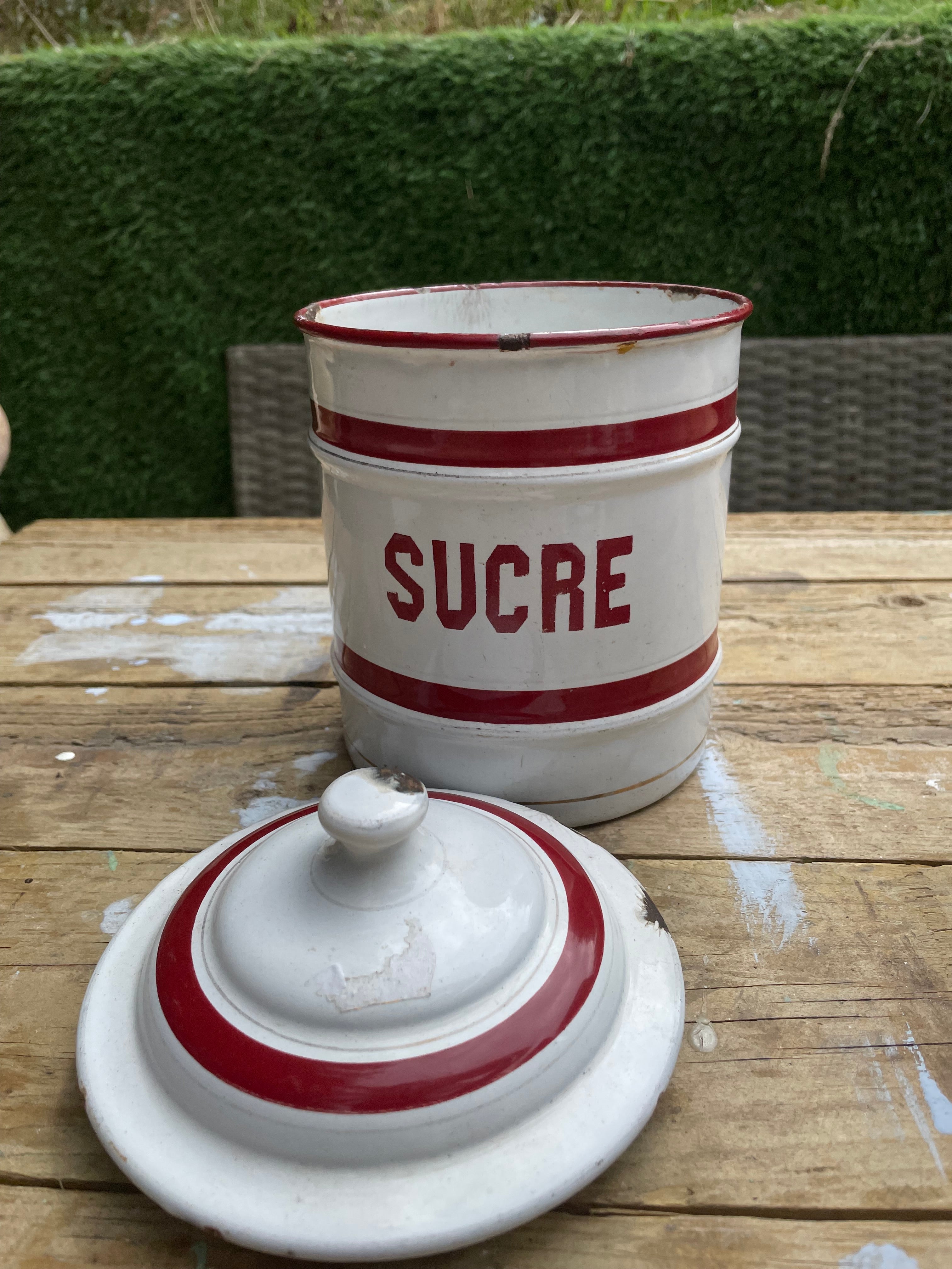 A vintage white enamel sugar canister with red stripes and the word 'SUCRE' printed in red, shown with its matching lid on a wooden table outdoors.
