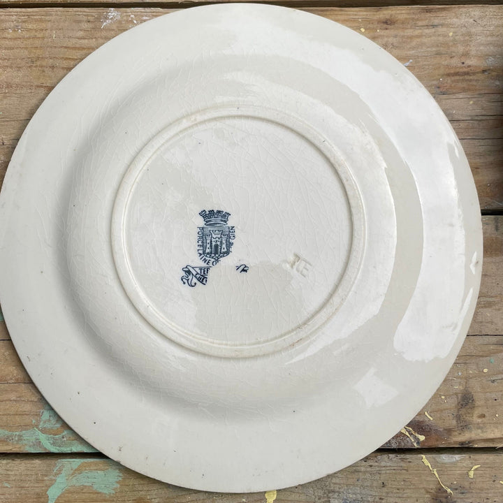 An off-white ceramic plate with a small blue monogram logo in the center, photographed from directly above on a rustic, weathered wooden plank surface.