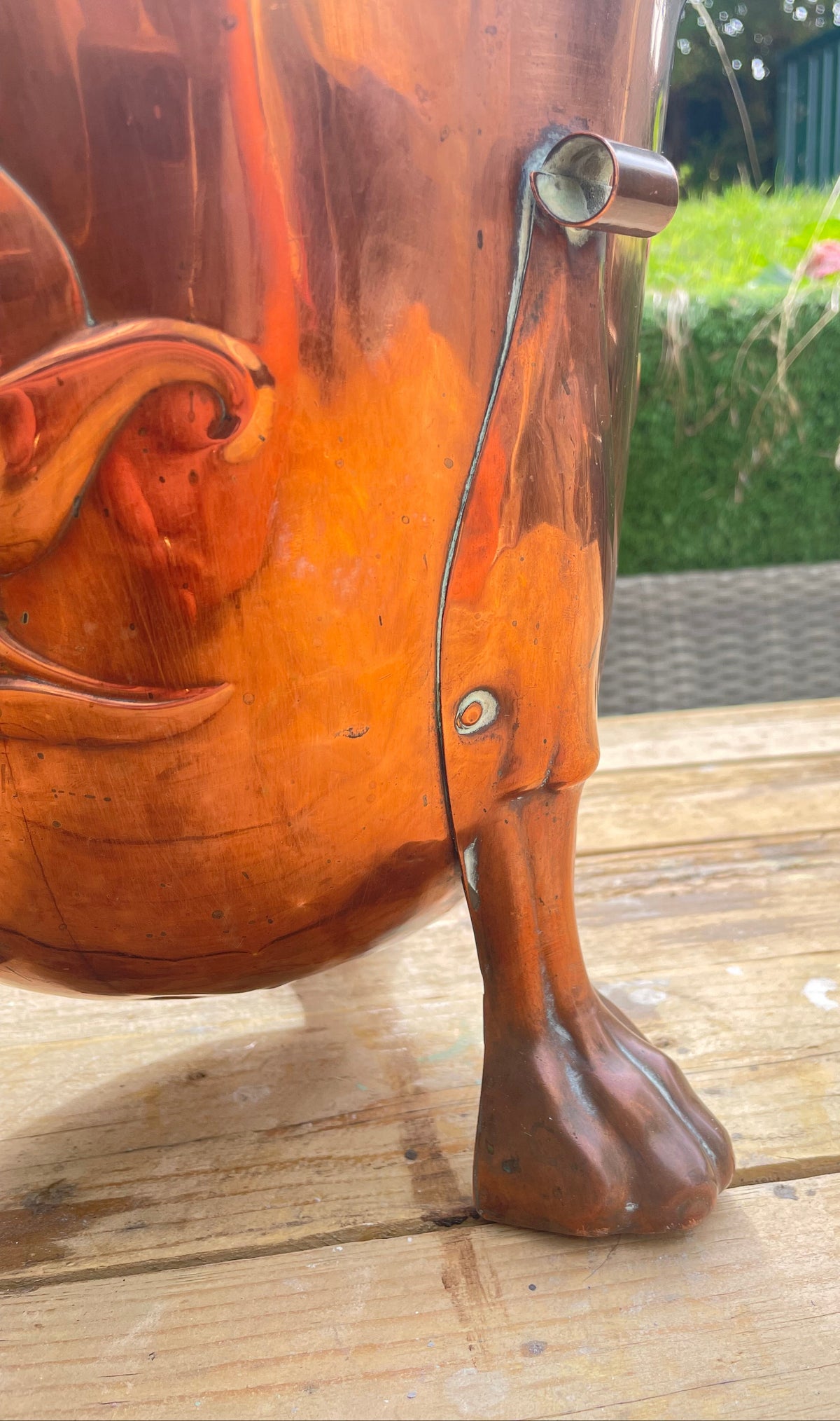 A close-up of a polished copper-colored metal container or vessel with a sculpted leg, resting on a wooden deck with a blurred garden background.