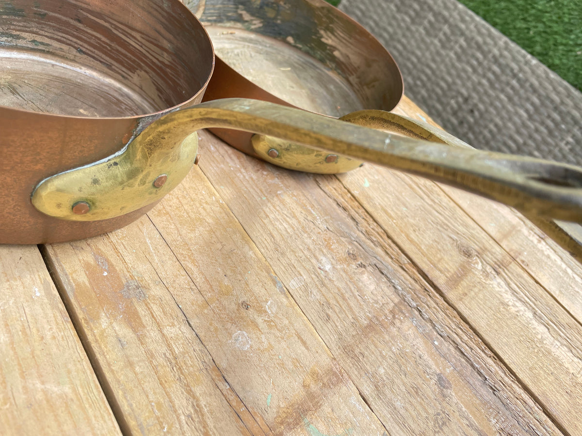 A close-up of a copper saucepan with a brass handle resting on a weathered wooden deck, with another pan partially visible behind it.