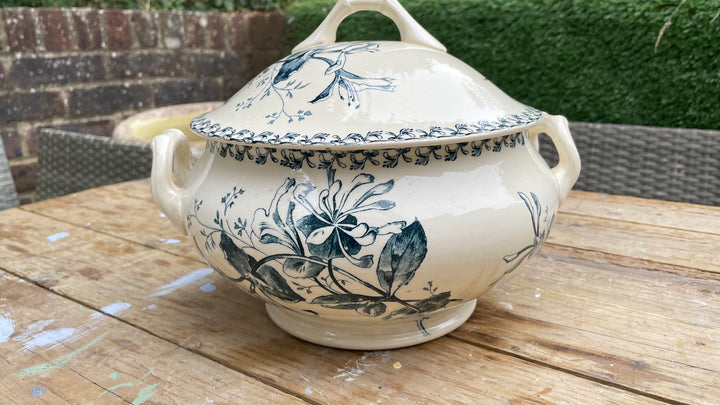 A cream-colored ceramic lidded tureen with blue floral illustrations, resting on a wooden table outdoors.
