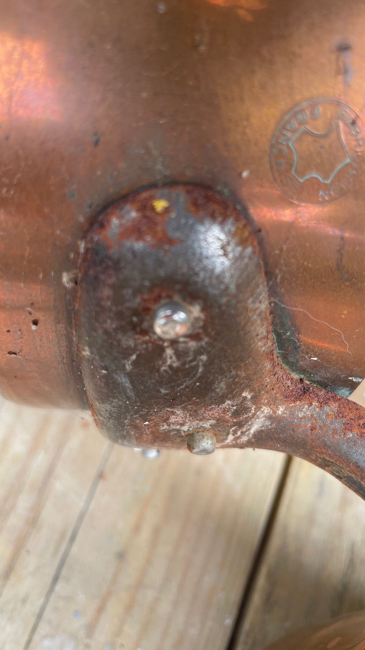 Close-up of a copper pot or vessel showing a weathered, rusted metal seam and rivet, with a stamped brand logo visible on the copper surface.
