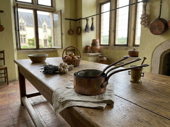 rustic kitchen scene with a wooden table holding a stack of copper pots on a cloth, along with garlic, herbs, a basket of apples, and hanging copper cookware in a warm yellow-walled room.