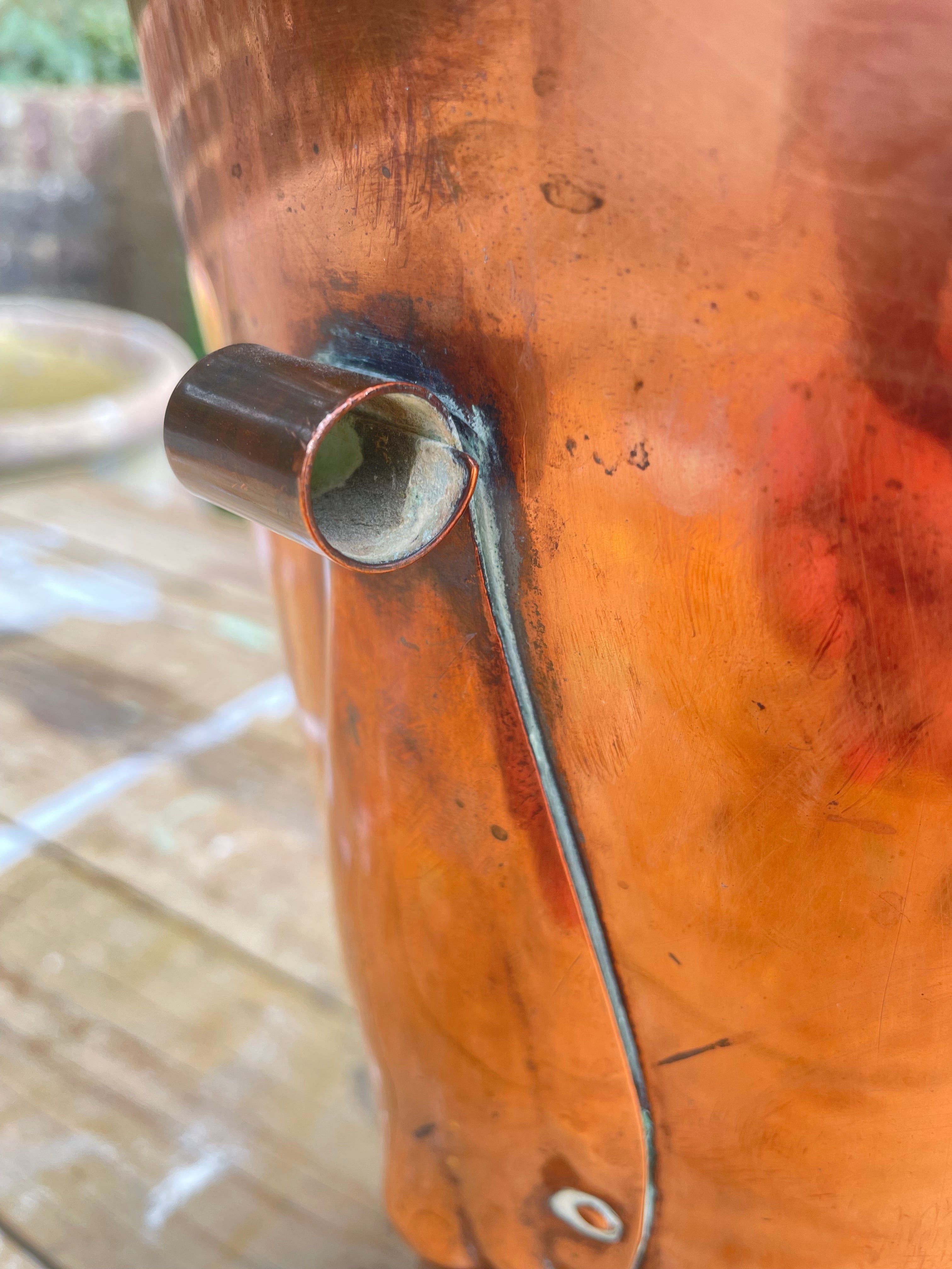 Close-up of a copper still pot with a copper pipe connection and a copper pipe running down the side, set on a wooden surface.