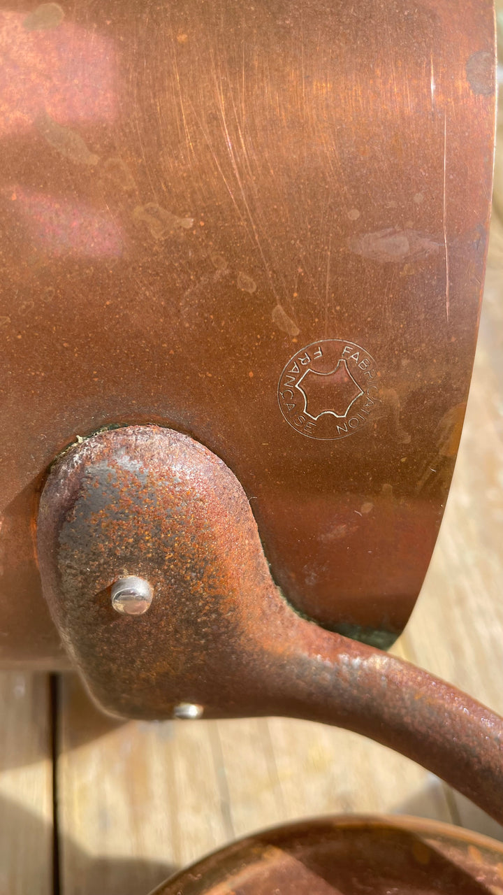 Close-up of a copper pot or pan with a rusted handle, showing a circular stamped maker's mark on the metal surface.