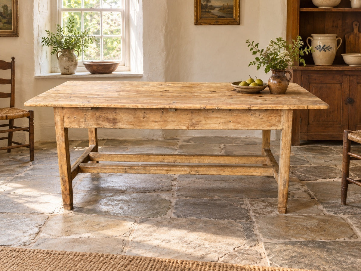 A rustic wooden dining table with a weathered, light brown finish and a lower stretcher bar, set in a bright, sunlit room with stone floors and potted plants.