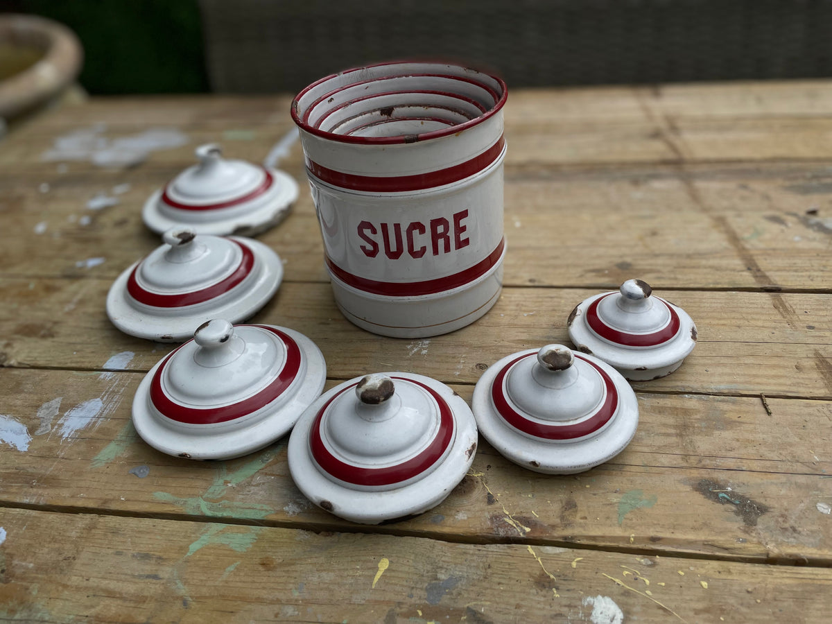 vintage white enamel sugar canister with red stripes and the word 'SUCRE' printed on the front, displayed on a rustic wooden table alongside five matching lids.