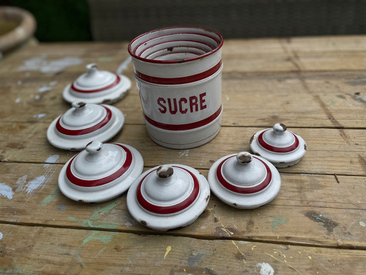 vintage white enamel sugar canister with red stripes and the word 'SUCRE' printed on the front, displayed on a rustic wooden table alongside five matching lids.