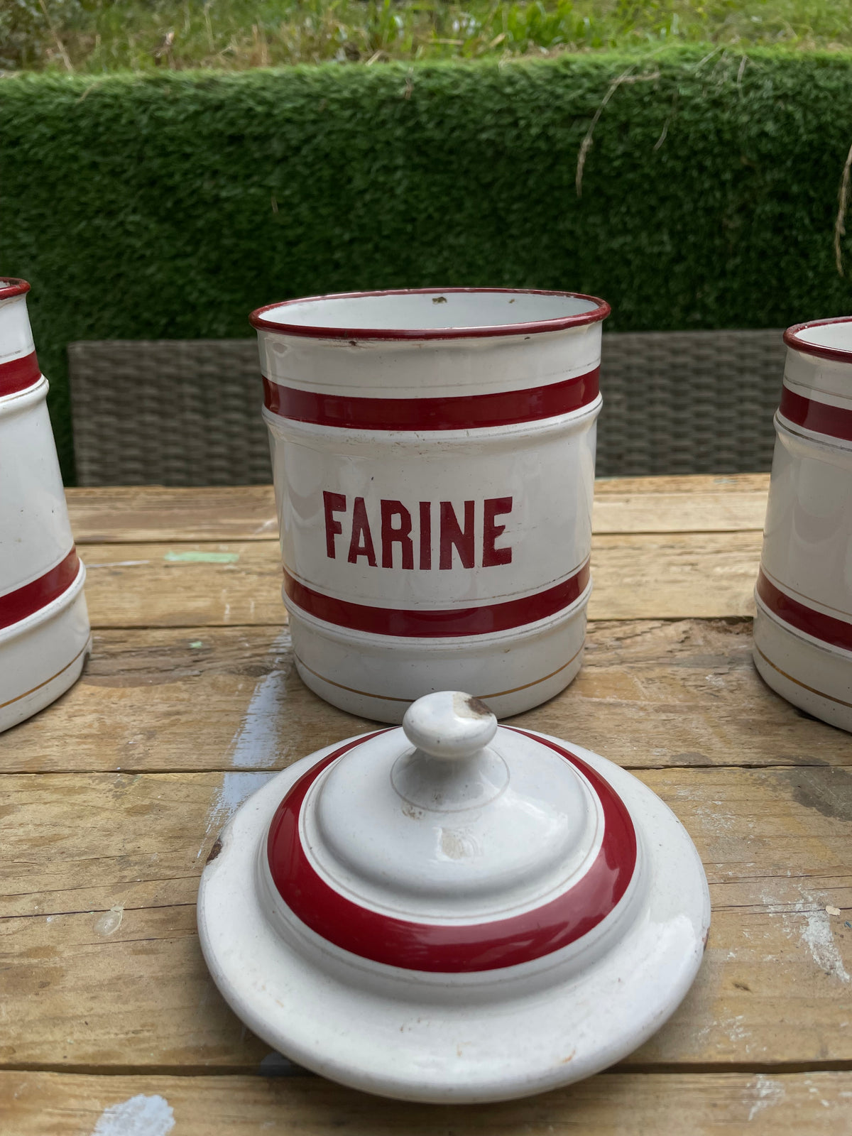 A white enamel canister with red stripes and the word 'FARINE' printed in red, shown on a wooden table outdoors with its matching lid in front. Other similar canisters are partially visible on both sides.