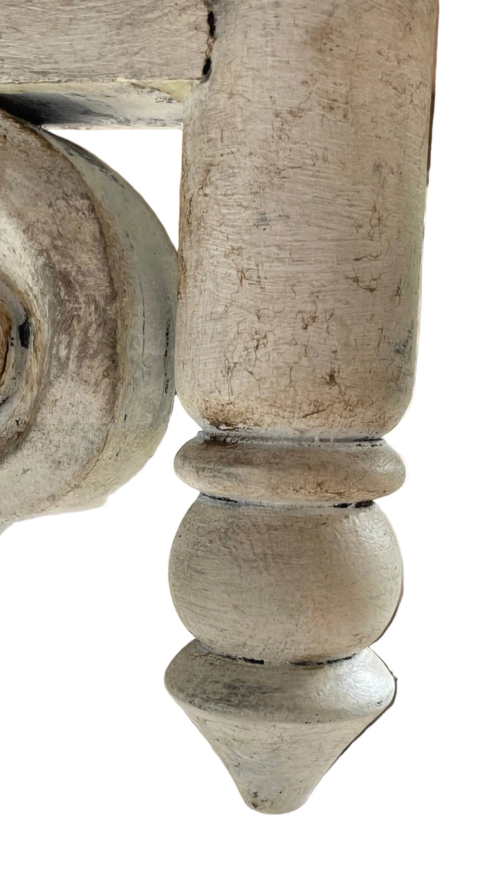 A close-up of a distressed, light beige painted wooden furniture leg with a turned ball and conical foot, showing weathered texture and chipped paint.