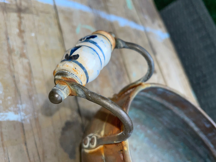Close-up of a vintage metal scoop or ladle with a white ceramic handle decorated with blue floral patterns and a rustic aged finish, resting on a weathered wooden surface.
